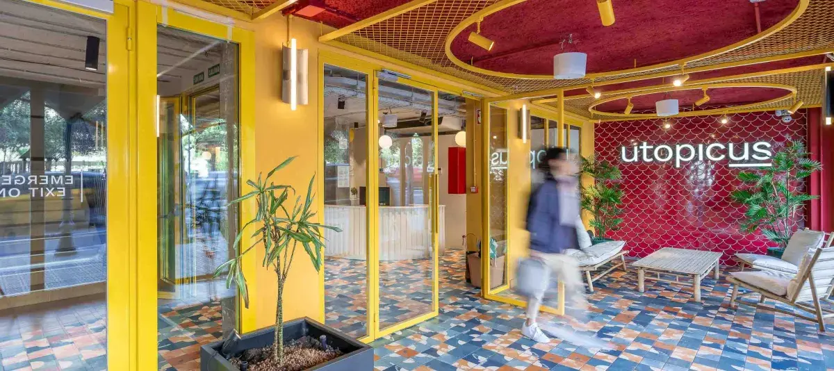 Una persona camina frente a una recepción con puertas amarillas, paredes rojas, plantas y suelo colorido.