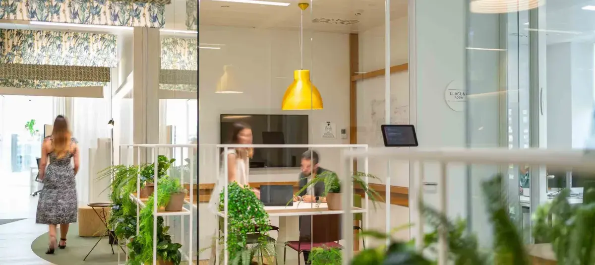 Una sala de reuniones con personas conversando, rodeada de plantas y decorada con lámpara amarilla, en una oficina moderna y luminosa.