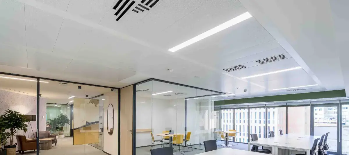 Mesas blancas vacías con sillas de oficina están en una sala amplia, dividida por paredes de vidrio transparentes, en un edificio moderno con grandes ventanas.