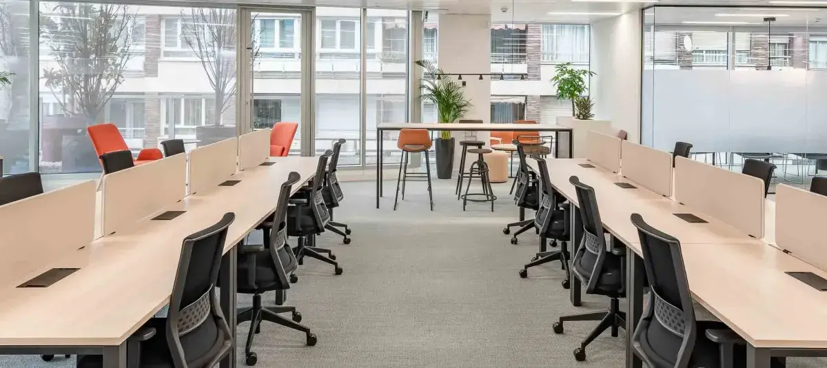 Escritorios con sillas en una oficina moderna; al fondo, mesas altas y plantas, junto a ventanas grandes que muestran el exterior urbano.
