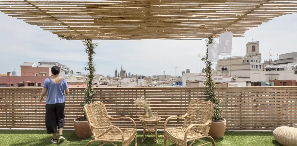 Una persona observa desde una terraza con pérgola de bambú y muebles de mimbre. Se ve una ciudad al fondo bajo un cielo despejado.