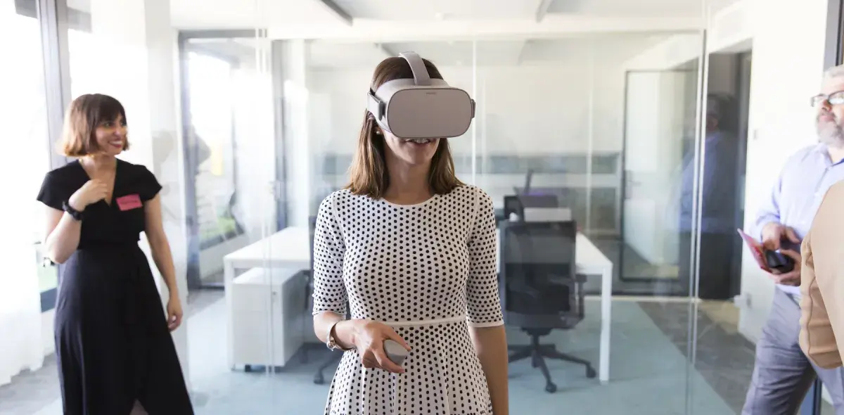 Una mujer usa gafas de realidad virtual en una oficina moderna con paredes de vidrio, mientras otras personas observan.