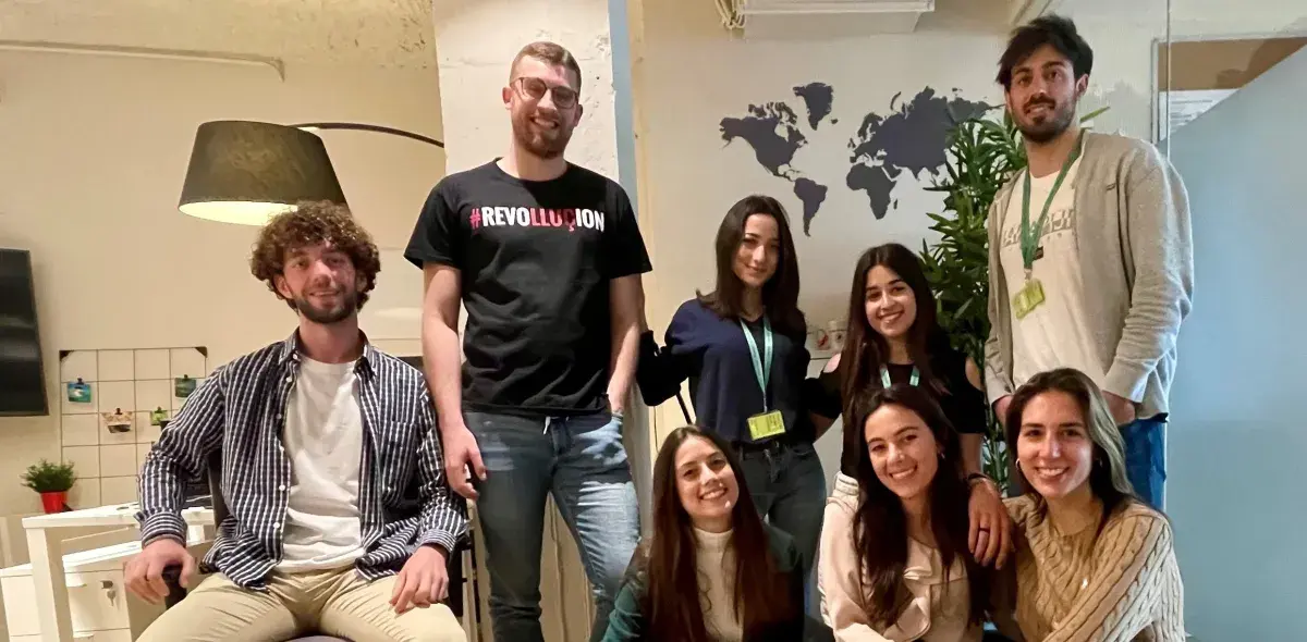 Un grupo de ocho personas sonríe a la cámara; algunos están sentados y otros de pie. Detrás hay un mapa del mundo en una pared y una planta. Una persona viste una camiseta con el texto "#REVOLUTION".