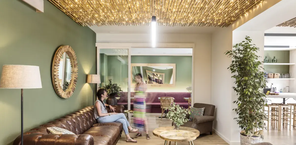 Una persona sentada en un sofá marrón interactúa con otra de pie frente a un espejo decorativo. La habitación tiene paredes verdes, iluminación cálida y plantas decorativas.