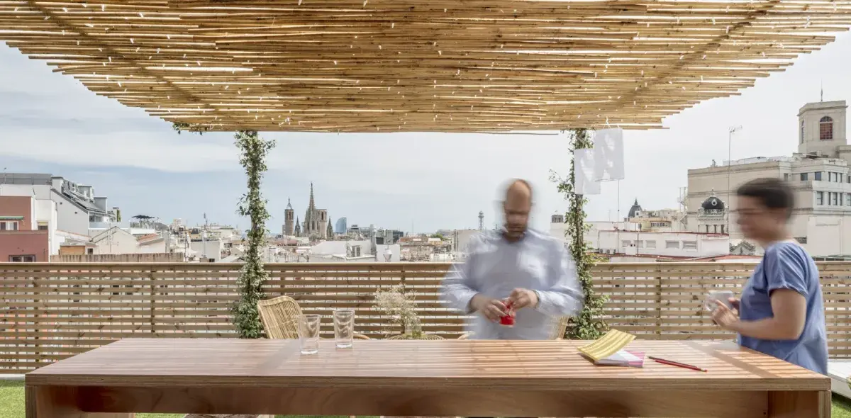 Una mesa de madera sostiene vasos vacíos mientras dos personas se mueven rápidamente bajo un techo de bambú en una terraza, con edificios urbanos al fondo.