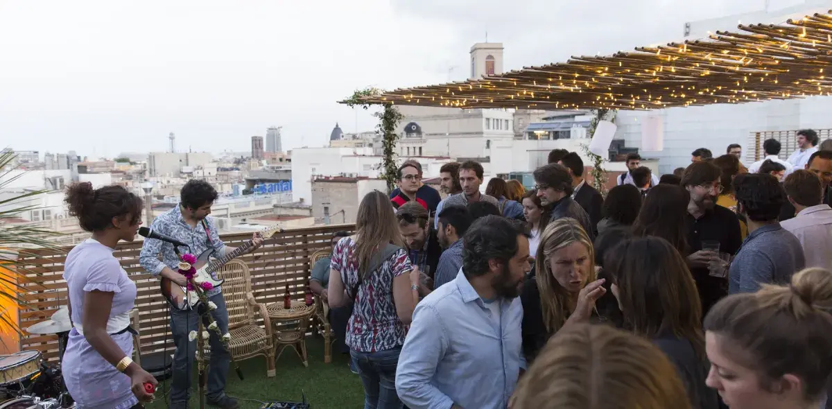 Varias personas socializan en una terraza decorada con luces, con vistas a la ciudad en el fondo.