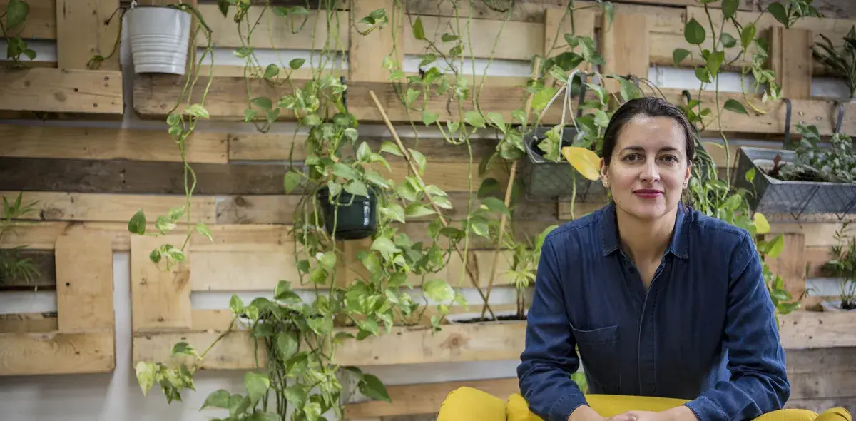 Una persona sentada en un espacio interior, rodeada de un fondo de palets de madera con varias plantas colgantes. Lleva una camisa azul oscuro.