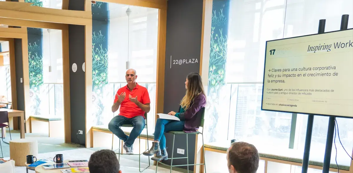 Hombre y mujer sentados conversan en una sala moderna con ventanas; pantalla muestra: "Inspiring Work, Claves para una cultura corporativa feliz y su impacto en el crecimiento de la empresa, Con Jaume Gurt, uno de los influencers más destacados de nuestro país y antiguo director de InfoJobs, Apúntate en: utopicus.es/agenda".