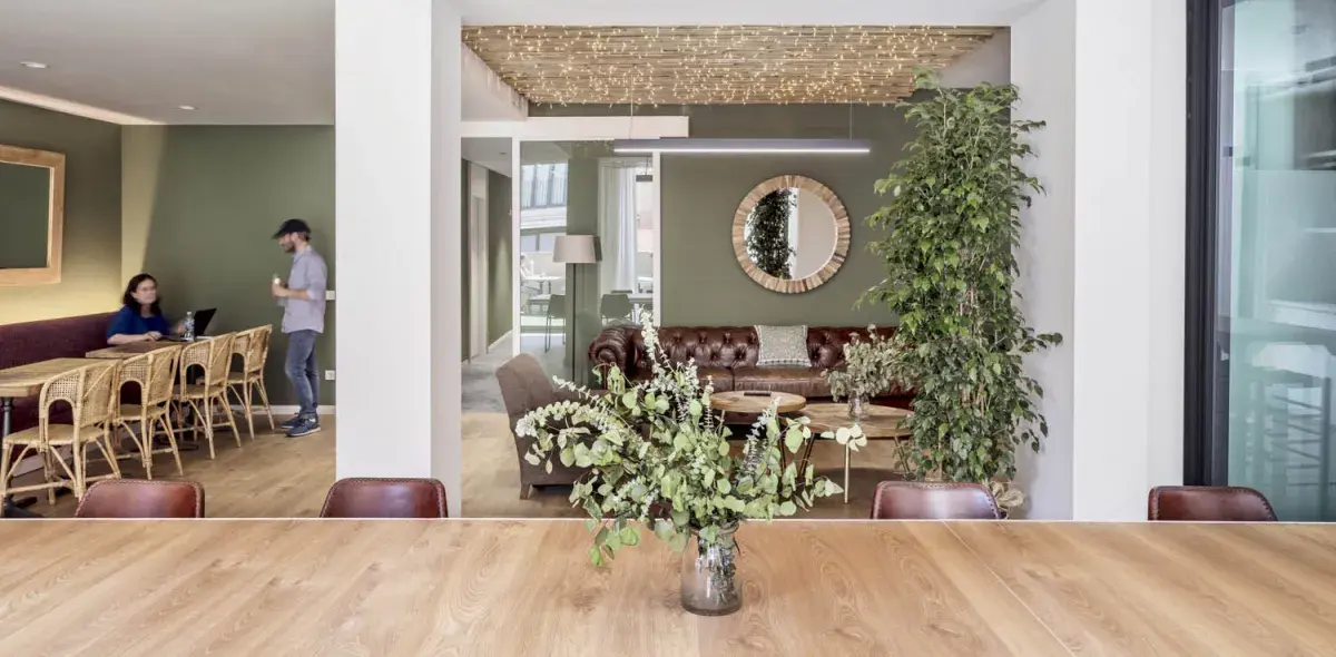Una mesa de madera sostiene un florero con hojas verdes, ubicada en una sala decorada con sofás, espejos y plantas. Al fondo, personas conversan junto a mesas y sillas de mimbre. Ambiente moderno y acogedor.