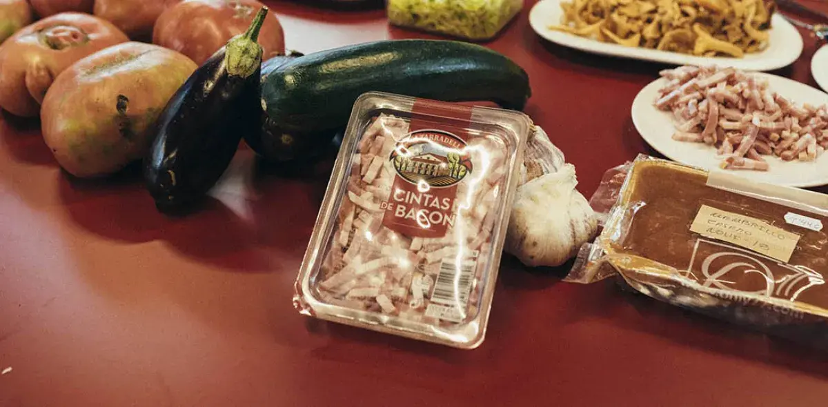 Sobre una mesa roja, un paquete de "cintas de bacon" está rodeado de calabacines, berenjenas, tomates, ajo y platos con alimentos variados, creando un ambiente culinario.