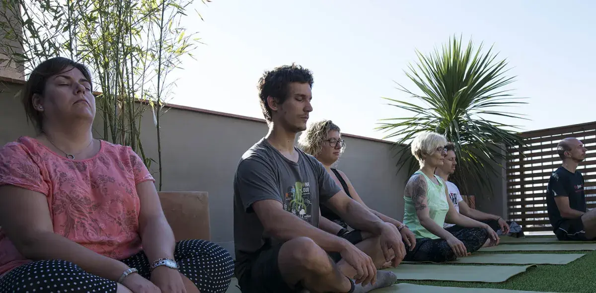 Personas meditando sentadas sobre colchonetas verdes, en una terraza rodeada de plantas y con cielo despejado.