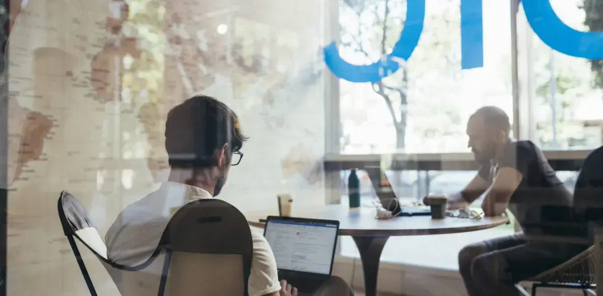 Personas trabajando en laptops, sentadas alrededor de una mesa redonda, dentro de una oficina con ventanas grandes. Letras azules parcialmente visibles en el vidrio.