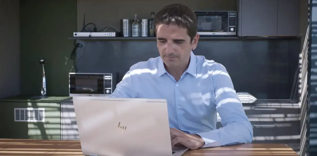 Un hombre usando una computadora portátil está sentado en una cocina, iluminado por luz que entra a través de persianas. Alrededor, se ven electrodomésticos como microondas y estantes con botellas.