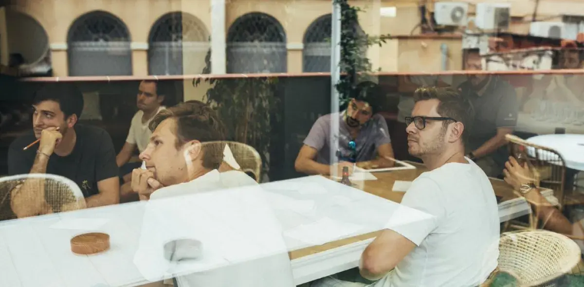 Varias personas están sentadas en una mesa, mirando atentamente en la misma dirección, en un ambiente interior con ventanas grandes.