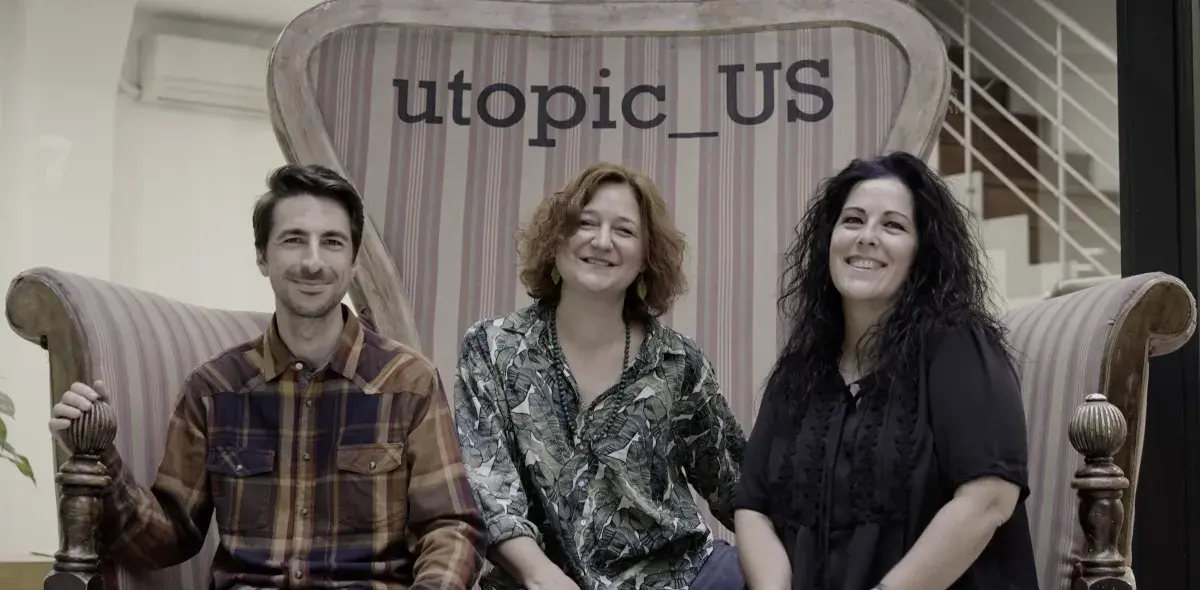 Tres personas sentadas en un sofá grande, sonriendo; detrás, el texto "utopic_US". Ambiente interior con escaleras al fondo.