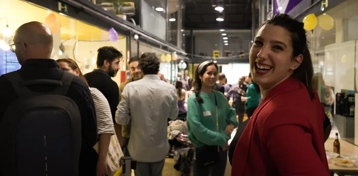 Persona sonriendo, vistiendo chaqueta roja, en un evento social concurrido con personas conversando en un espacio cerrado decorado con globos.