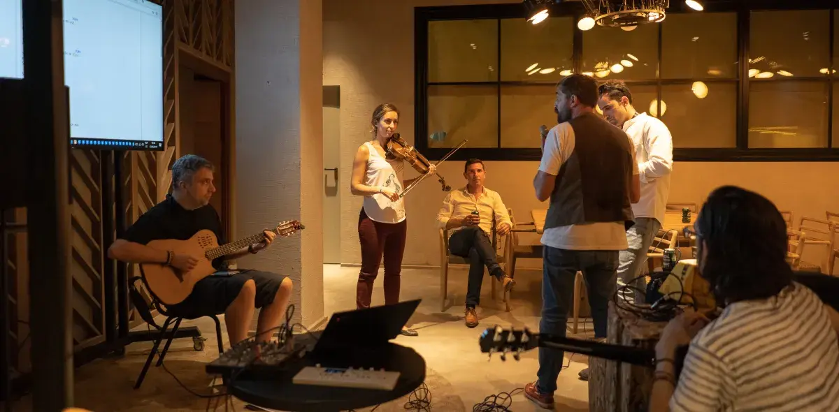 Un grupo de personas toca instrumentos musicales, como la guitarra y el violín, en una sala iluminada. Algunos están sentados, otros de pie, rodeados de equipos electrónicos.