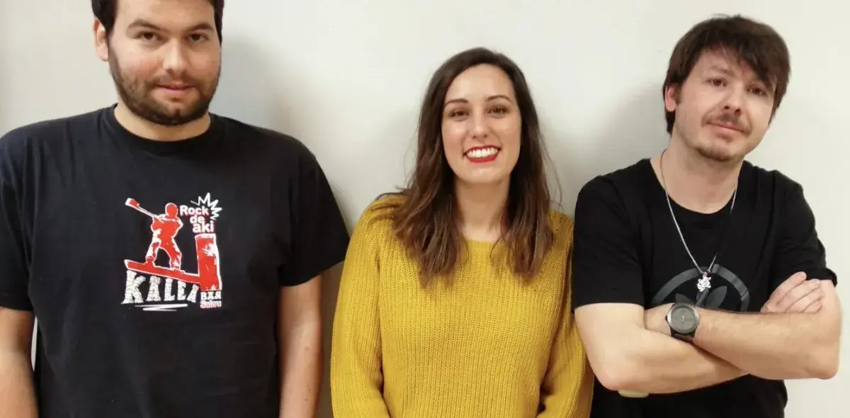 Tres personas de pie, sonriendo frente a una pared blanca. Una lleva camiseta negra con texto: "Rock de aki Kalla".