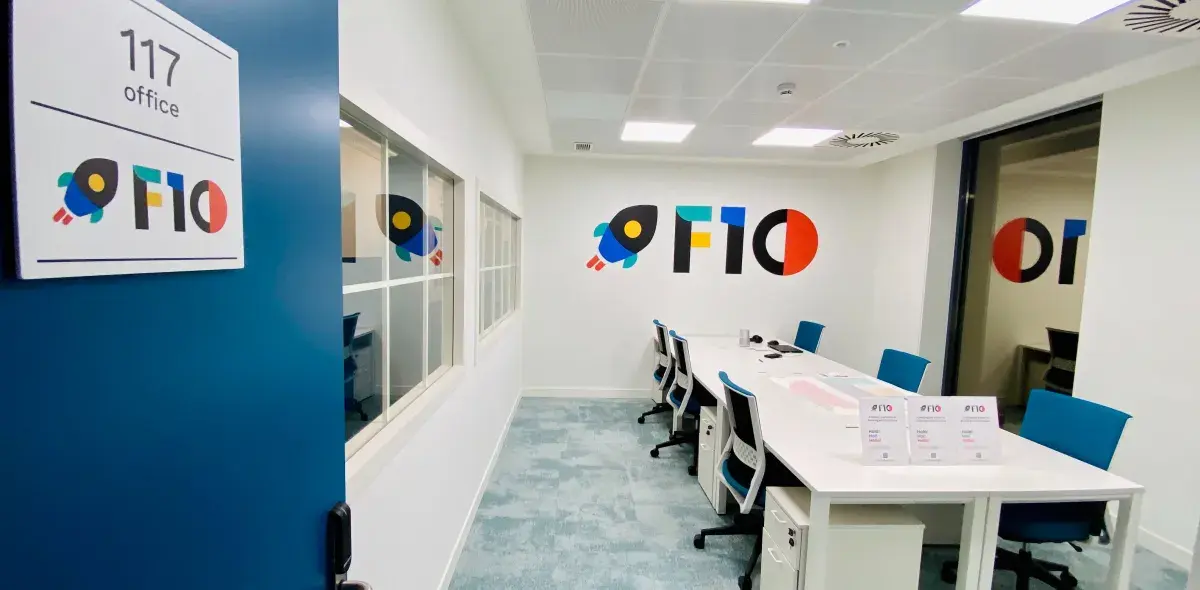Una sala de reuniones moderna con seis sillas azules alrededor de una mesa blanca. En la pared, el texto "F10" y un dibujo de cohete. La puerta tiene un cartel: "117 office".