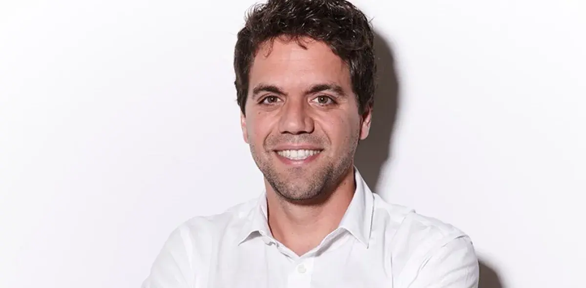 Un hombre sonriente lleva una camisa blanca, de brazos cruzados, sobre un fondo blanco liso.