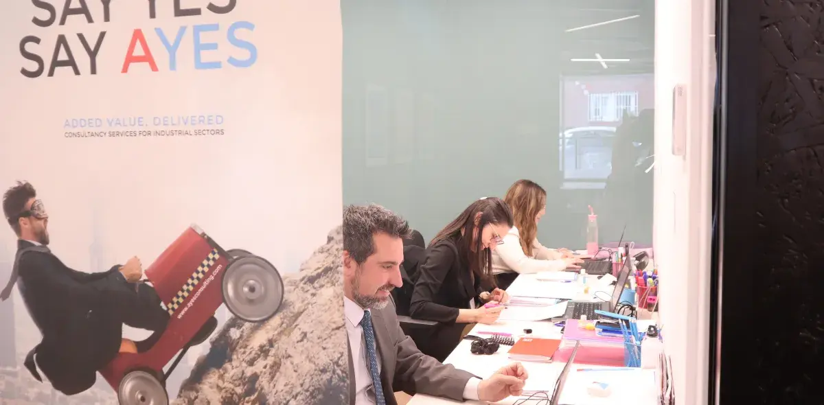 Varias personas trabajando en un escritorio en una oficina moderna. Cartel publicitario dice "SAY YES SAY AYES" y "ADDED VALUE, DELIVERED".