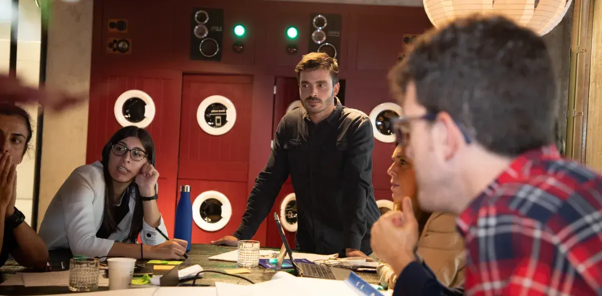 Varias personas conversan alrededor de una mesa en un ambiente interior con paredes rojas decoradas con círculos.