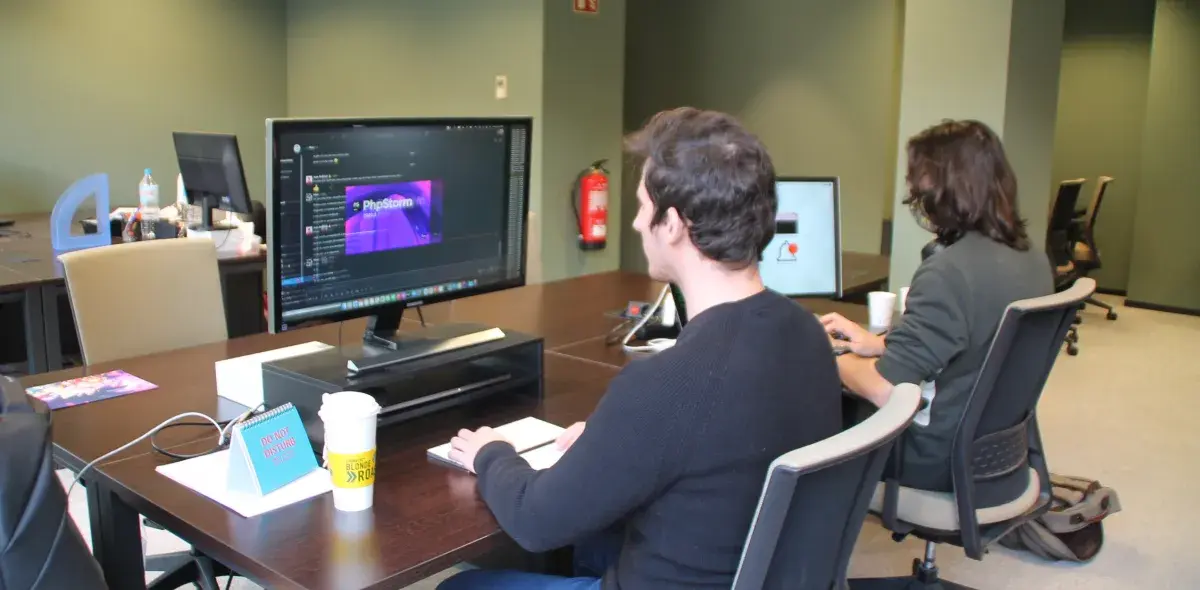 Un hombre trabaja en una computadora con un editor de texto abierto, en una oficina compartida. Sobre la mesa, un cartel dice "DO NOT DISTURB" y una taza "BLONDE ROAST".