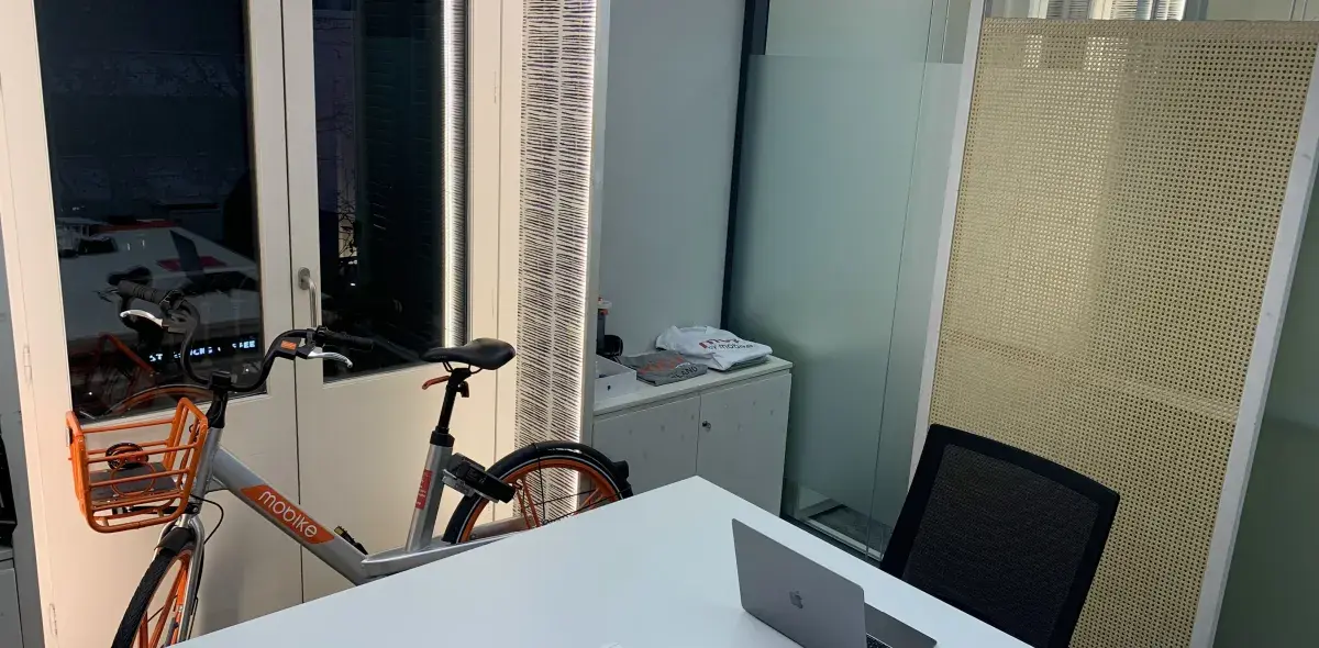 Una bicicleta estática se encuentra al lado de una mesa blanca con una laptop abierta en una oficina moderna con paredes de vidrio y muebles minimalistas.