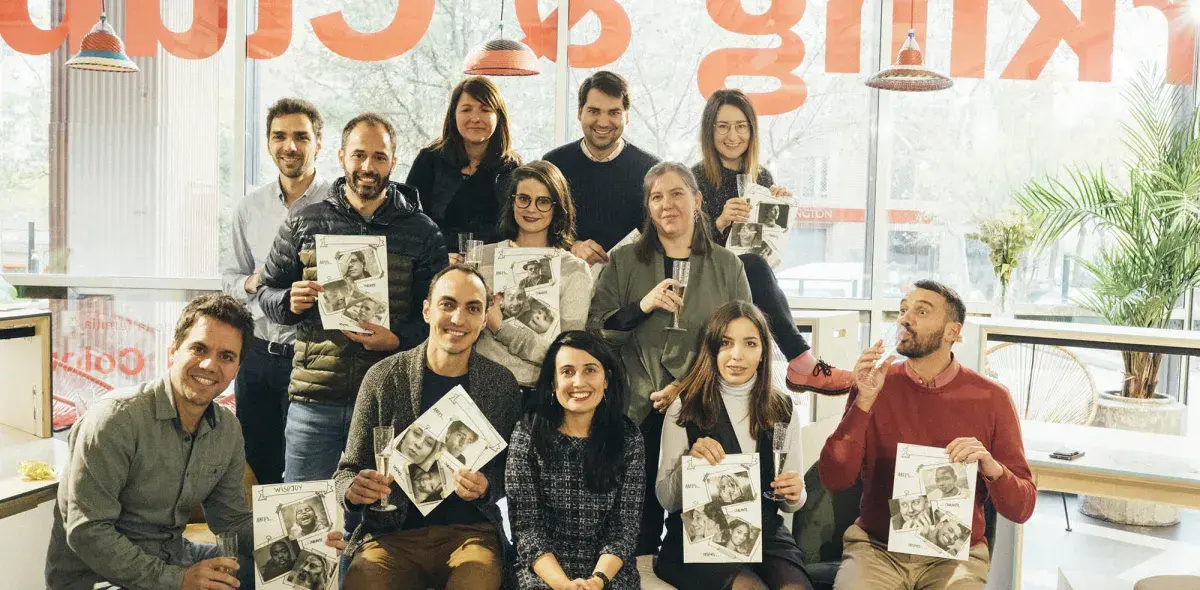 Un grupo de personas sostiene revistas sonrientes en una cafetería luminosa y moderna. Al fondo, un cartel rojo parcialmente visible.