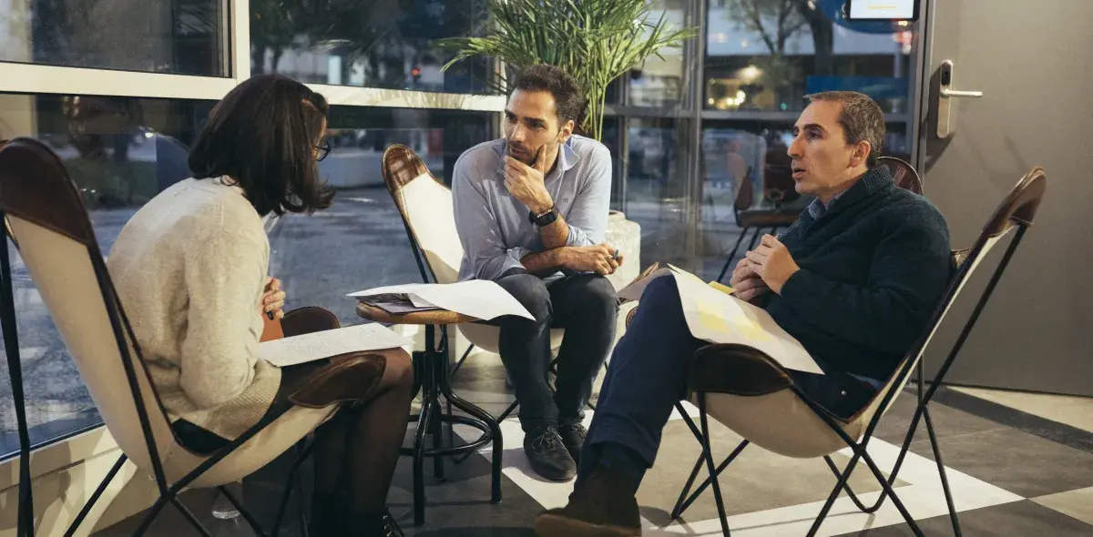 Tres personas sentadas en sillas, discutiendo documentos, en un ambiente interior junto a una ventana grande.