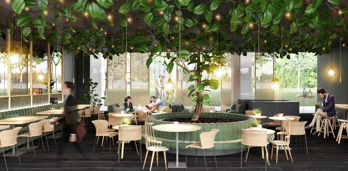 Un café moderno tiene mesas de madera y sillas, con un árbol central y luces colgantes. Grandes ventanales muestran un parque exterior, mientras personas conversan y caminan dentro del espacio.