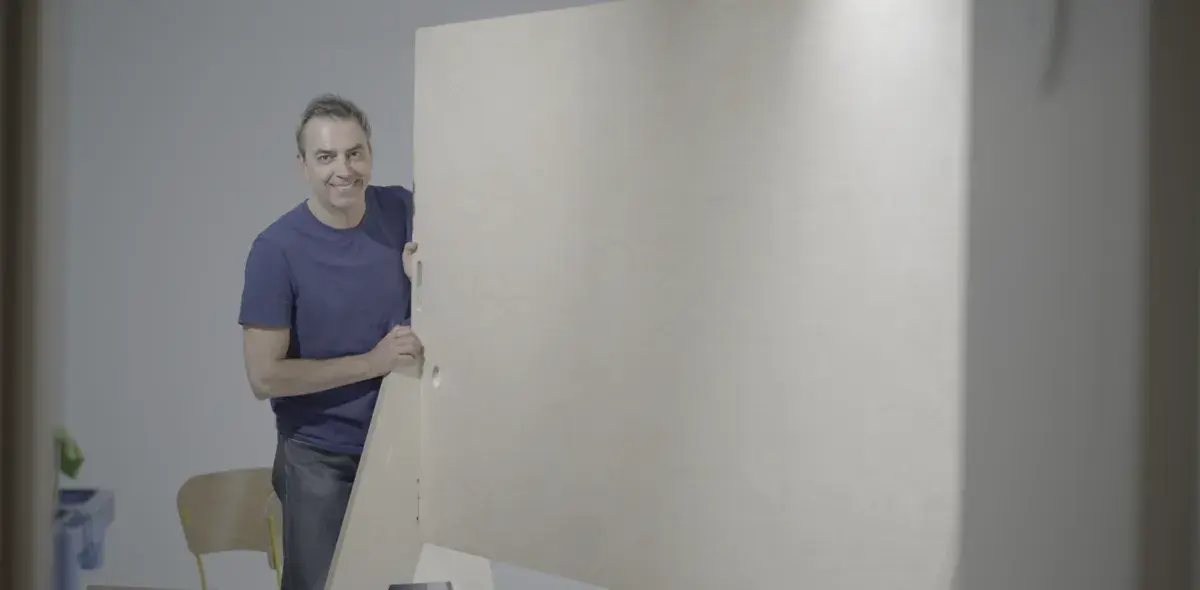 Un hombre, sonriendo, sostiene una puerta grande de madera en una habitación clara con una silla al fondo.