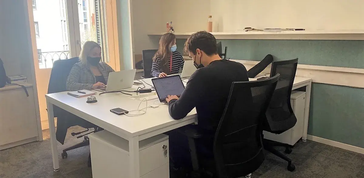 Tres personas trabajan en laptops, sentadas alrededor de una mesa blanca en una oficina iluminada por luz natural.