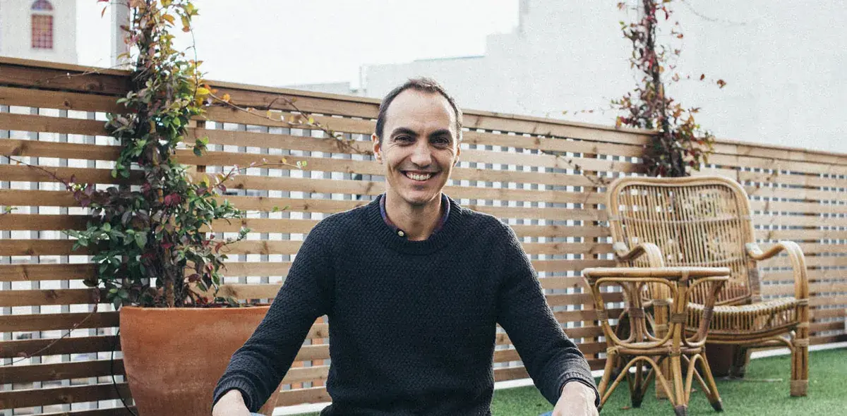 Un hombre sonriente está sentado en una terraza. A su alrededor, hay plantas y sillas de mimbre.