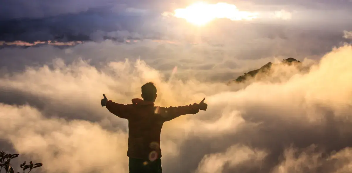 Una persona con brazos extendidos y pulgares levantados mira hacia un amanecer entre nubes, sobre una montaña.