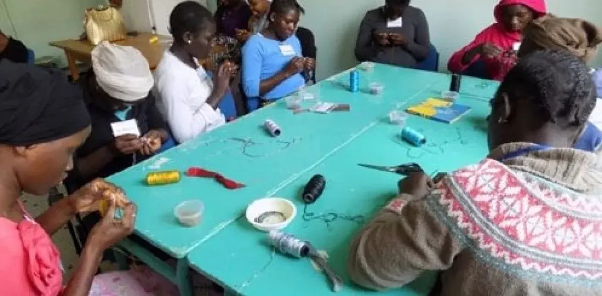 Un grupo de personas realiza manualidades con hilos en una mesa azul; hay bobinas y herramientas dispersas alrededor. Están concentrados y colaborando en un espacio interior.