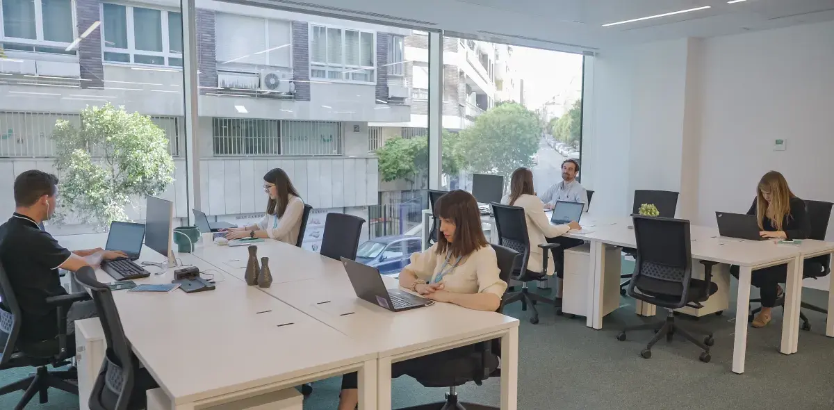 Personas trabajando en laptops. Están sentadas en escritorios blancos en una oficina iluminada con una gran ventana que muestra un edificio y árboles afuera.