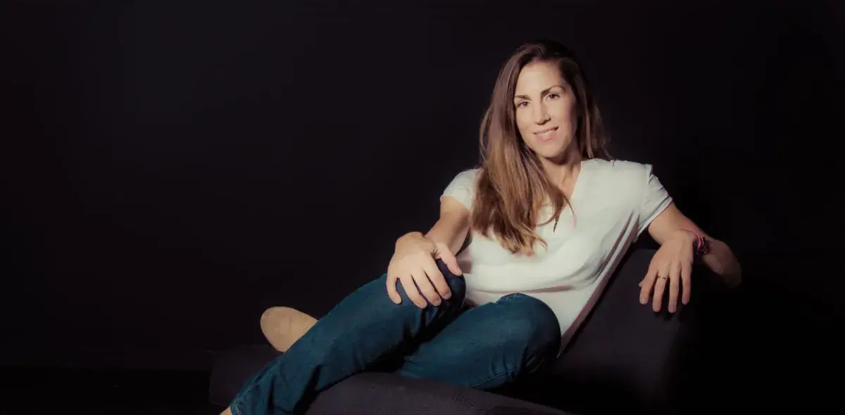 Una mujer está sentada relajadamente en un sillón negro, con un fondo oscuro, vistiendo una camiseta blanca y jeans.