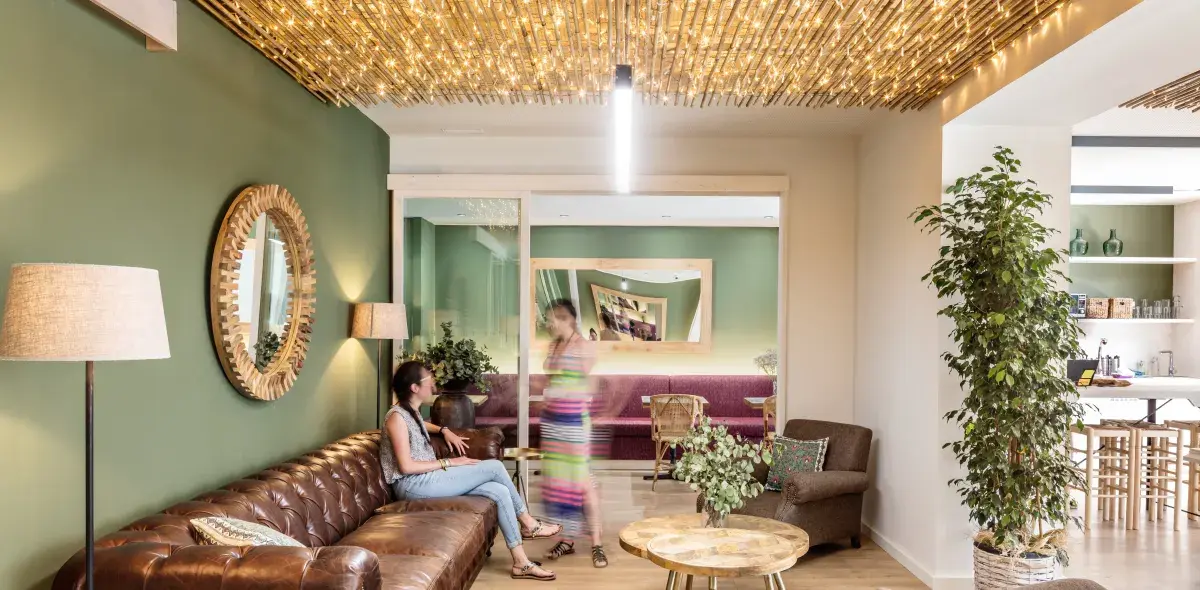 Una mujer está sentada en un sofá de cuero marrón, hablando con otra que pasa caminando. La habitación tiene paredes verdes, un techo con luces cálidas, espejos y plantas decorativas.