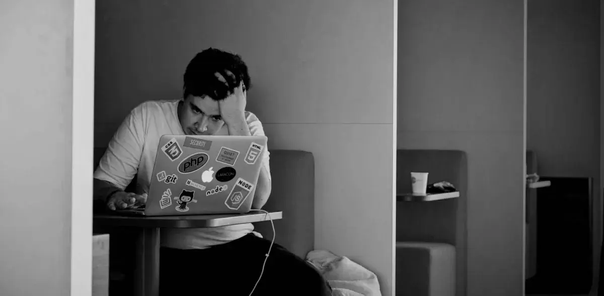 Persona concentrada usando una laptop llena de pegatinas, en una sala de estar, apoyando su cabeza con una mano y sosteniendo la otra en el teclado.