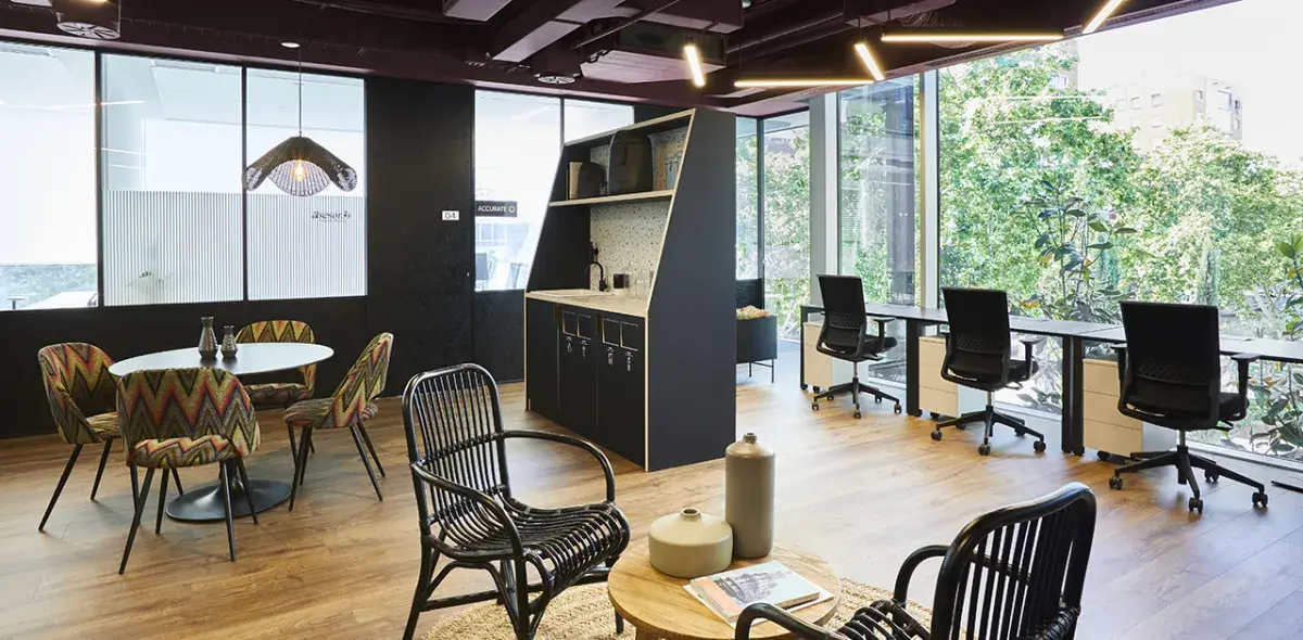 Espacio de oficina moderno; mesa y sillas en el centro, escritorios al fondo junto a ventanas grandes, con luz natural y vista a árboles. Lámparas colgantes iluminan el área.