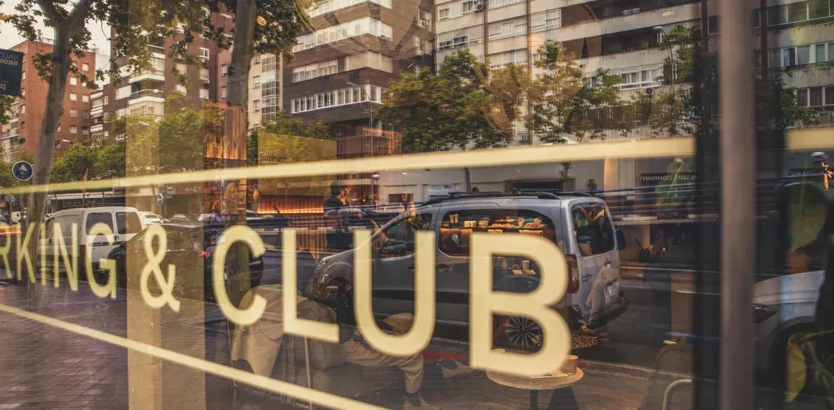 Reflejo de la calle en una ventana con letras grandes que dicen "WORKING & CLUB", rodeado de edificios y árboles.