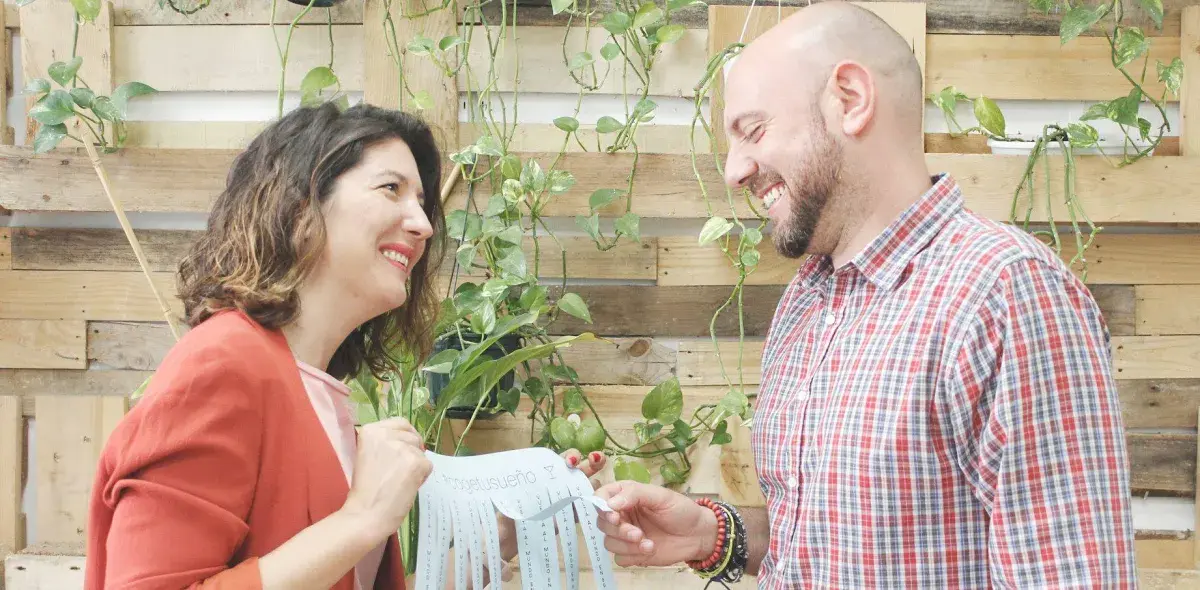 Una mujer y un hombre sonríen sosteniendo un papel con texto ilegible. Están en un entorno con palés y plantas verdes colgantes.