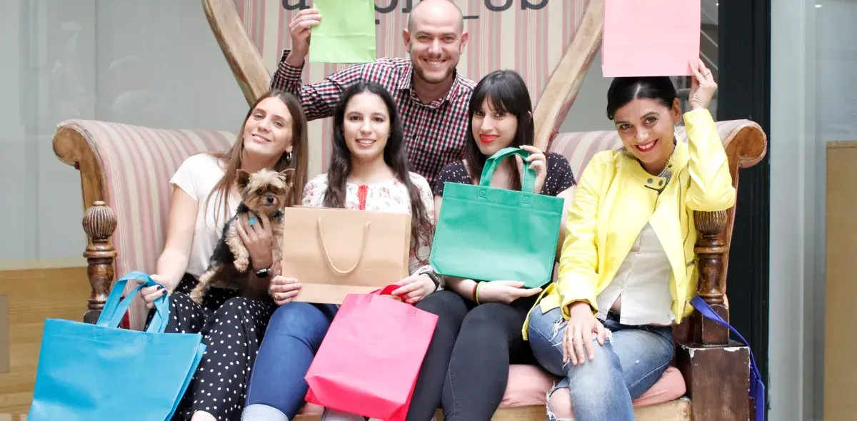 Varias personas sonrientes sostienen bolsas de compras de colores mientras están sentadas en un sofá estilizado en un entorno interior. Una persona sostiene un perro pequeño.