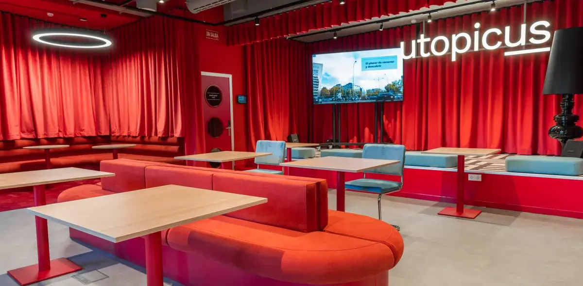Sofás rojos y mesas vacías en un salón con cortinas rojas, pantalla grande y el texto "utopicus".