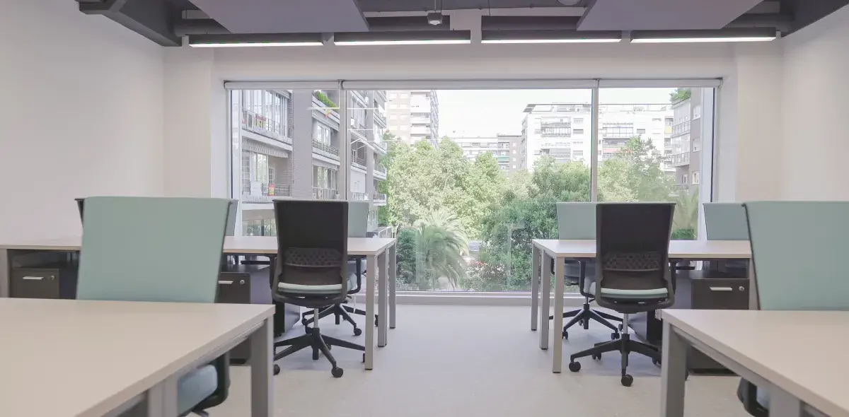 Escritorios vacíos con sillas giratorias están frente a una ventana grande que muestra edificios y árboles afuera.