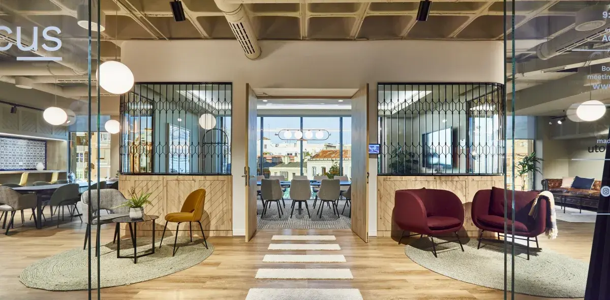 Una moderna sala de reuniones con mesas y sillas, iluminación colgante, y puertas abiertas hacia el exterior urbano. La pared de cristal muestra reflejos, añadiendo elegancia al ambiente.