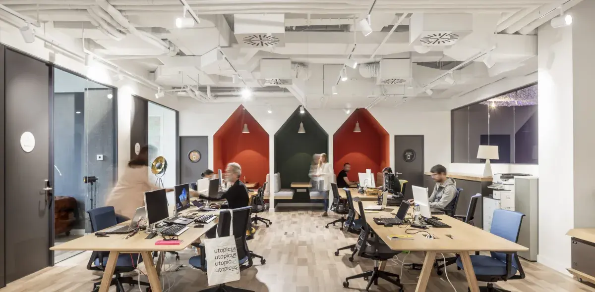 Varias personas trabajan en sus computadoras en una oficina moderna, con cabinas de privacidad coloridas y decoración minimalista.