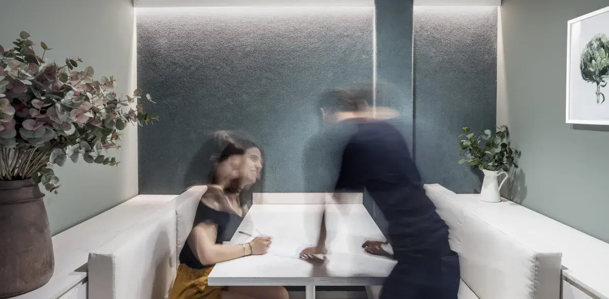 Una mujer sentada en un banco mezcla observa a un hombre desenfocado colocarse en la mesa en un moderno comedor con paredes verdes y plantas decorativas.