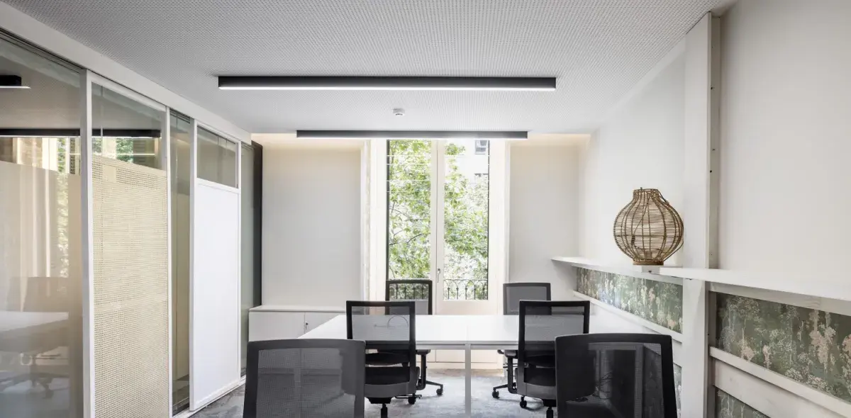 Mesa con sillas rodeándola, situada en una oficina con ventanas grandes y una lámpara decorativa sobre una repisa.paredes blancas.